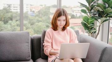 mooie aziatische vrouw die computer of laptop gebruikt terwijl ze op de bank in haar woonkamer ligt. gelukkige vrouw die thuis online winkelen koopt. levensstijl vrouw thuis concept. foto
