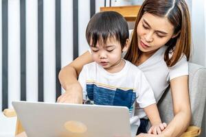 jong Aziatisch moeder haar weinig aanbiddelijk zoon gebruik makend van laptop op zoek Bij computer scherm genieten van aan het kijken grappig sociaal media aan het doen online boodschappen doen ontspannende Aan sofa Bij huis samen foto