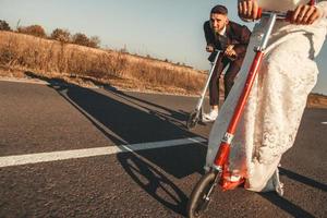 lachend bruidspaar rijden op scooters langs de weg buiten de stad bij zonsondergang. plaats voor tekst of reclame foto