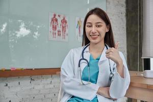 portret van mooie vrouwelijke arts van Aziatische etniciteit in uniform met stethoscoop, duim omhoog, glimlachend en kijkend naar de camera in een kliniek, een persoon die expertise heeft in professionele behandeling. foto