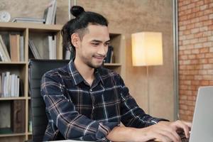 jonge mannelijke werknemer van aziatische etniciteit gebruikt laptop om creatief werk te doen op een wit bureau voor een boekenplank van een informele werkplek, startende ondernemer en online e-commerceberoep. foto