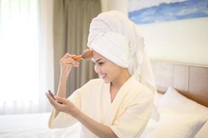 aantrekkelijke gelukkige vrouw in witte badjas past natuurlijke make-up toe met cosmetische poederborstel, schoonheidsconcept. foto