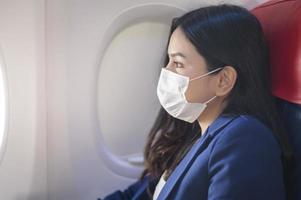 een jonge zakenvrouw draagt een beschermend masker aan boord van het vliegtuig, reist onder covid-19 pandemie, veiligheidsreizen, protocol voor sociale afstand, nieuw normaal reisconcept