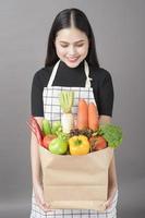 portret van mooie jonge vrouw met groenten in boodschappentas in studio grijze achtergrond