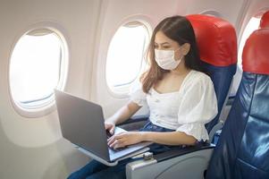 een jonge vrouw met een gezichtsmasker gebruikt laptop aan boord, nieuw normaal reizen na covid-19 pandemisch concept foto