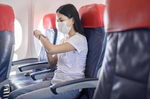 reizende vrouw met een beschermend masker aan boord in het vliegtuig met behulp van een slim horloge, reizen onder covid-19 pandemie, veiligheidsreizen, protocol voor sociale afstand, nieuw normaal reisconcept foto
