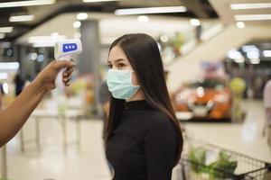 close-up hand met temperatuurpistool met jonge vrouw, sociale afstandsmaatregel voor covid-19-preventie in winkelcentrum foto