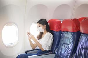 een jonge vrouw met gezichtsmasker gebruikt smartphone aan boord, nieuw normaal reizen na covid-19 pandemisch concept