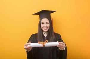 portret van gelukkige mooie vrouw in afstudeerjurk houdt onderwijscertificaat op gele achtergrond foto