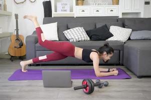 een vrouw doet yogaplank en kijkt naar online trainingshandleidingen op haar laptop in de woonkamer, fitnesstraining thuis, concept voor gezondheidszorgtechnologie. foto