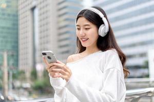 jong Aziatisch meisje met een koptelefoon die naar muziek luistert vanaf een mobiele telefoon tegen de achtergrond van het bouwen van een stad foto
