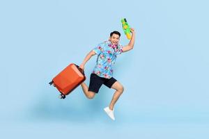 springende knappe aziatische toeristenmens die met waterpistool en bagage reist tijdens songkran festival studio schoot blauwe achtergrond foto