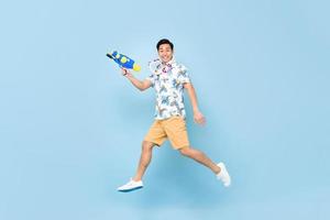 knappe glimlachende jonge aziatische man spelen met waterpistool en springen in studio blauwe achtergrond voor songkran festival in thailand en zuidoost-azië foto