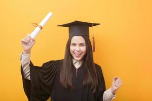 portret van gelukkige mooie vrouw in afstudeerjurk houdt onderwijscertificaat op gele achtergrond foto