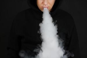 de man rookt een elektronische sigaret op de donkere achtergrond, mannen in hoody vaping en laat een wolk van damp vrij. man met vapen op zwarte achtergrond foto