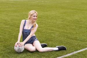 blond meisje met een bal zittend op een voetbalveld. foto