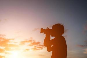 silhouet van een jonge man met camera, strek de armen uit terwijl zonsondergang. foto