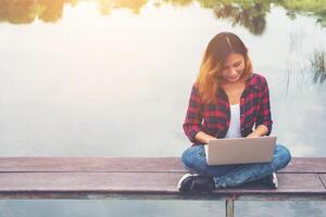 jonge gelukkige hipster vrouw die werkt met haar laptop zittend op de pier, ontspannen genieten met de natuur. foto