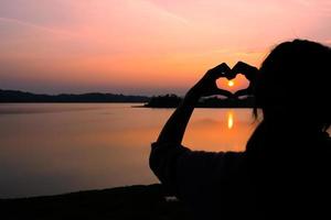 silhouet van de hand van de vrouw die zich in hartvorm vormt rond de zon bij zonsondergang in de buurt van het meer foto