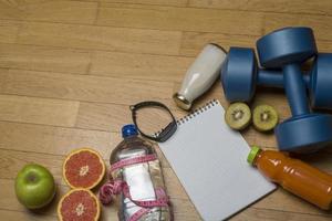 training, oefening, opgewektheid en gezondheid - twee plastic dumbbells, een notitieboekje, mineraalwater met sap, fruit en een pen op de houten vloer. foto