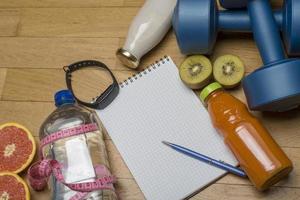 training, oefening, opgewektheid en gezondheid - twee plastic dumbbells, een notitieboekje, mineraalwater met sap, fruit en een pen op de houten vloer. foto