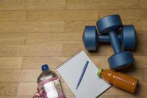 training, oefening, opgewektheid en gezondheid - twee plastic dumbbells, een notitieboekje, mineraalwater met sap en een pen op de houten vloer. foto