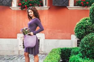 vrouw, gekleed in paarse polka dot jurk, met gebreide tas met bloemen, poserend in de straat van de stad. kopiëren, lege ruimte voor tekst foto