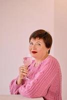 vrolijke rijpe vrouw in roze trui drinken van rose wijn en glimlachen. zelfvoorziening, vakantie, vieren concept foto