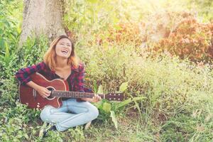 jonge mooie hipster vrouw gitaarspelen zittend op het gras in het park, ontspannen met de natuur. foto