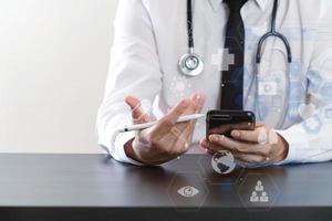 close-up van slimme arts die werkt met slimme telefoon en stethoscoop op donker houten bureau met virtueel pictogramdiagram foto