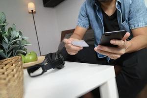 jonge man zit op een bank online te winkelen met een smartphone luistert muziek met de koptelefoon houdt thuis een creditcard op zijn hand, omni-kanaalconcept foto