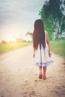 klein meisje met lang haar dat een jurk draagt, loopt van je weg op een landelijke weg. foto