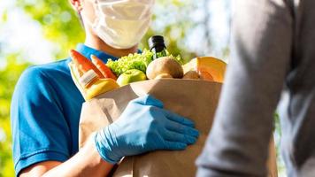 hygiënische bezorger die gezichtsmasker en rubberen handschoenen draagt terwijl hij boodschappen bij de klant thuis bezorgt, voedselbezorging in de tijd van pandemisch concept foto