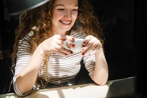Gelukkig lachende jonge blanke vrouw die koffie drinkt terwijl ze 's morgens vroeg thuis naar video kijkt op een laptopcomputer foto
