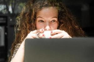 jonge blanke vrouw die koffie drinkt tijdens het kijken naar video op een laptop thuis in de vroege ochtend foto