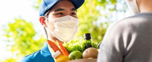 hygiënische aziatische bezorger die gezichtsmasker draagt terwijl hij boodschappen bij de klant thuis bezorgt, voedselbezorging in de tijd van pandemisch concept foto