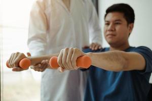 Aziatische fysiotherapeut die een patiënt helpt die dumbbells optilt om zijn herstel te doorlopen met gewichten in de kliniekkamer. foto