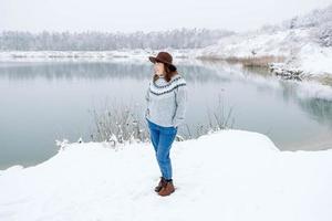 vrouw in een bruine hoed en trui staat aan de oevers van een met sneeuw bedekt meer en bos foto