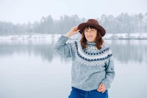 vrouw in een bruine hoed en trui staat aan de oevers van een met sneeuw bedekt meer en bos foto