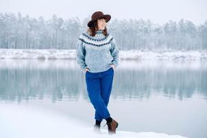 lachende vrouw in een bruine hoed en trui staat aan de oevers van een met sneeuw bedekt meer en bos foto