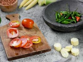 gesneden baby maïs, gesneden tomaten op een houten snijplank. cayennepeper in een vijzel. voedsel recept concept foto