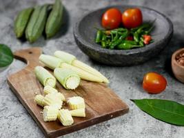 gesneden baby maïs op een houten snijplank. cayennepeper en tomaten in de vijzel. voedsel recept concept foto