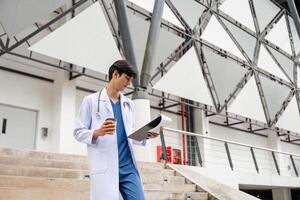 zelfverzekerd mannetje dokter staand buiten modern ziekenhuis gebouw met klembord en koffie kop foto