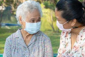verzorger helpt en praat met aziatische senior of oudere oude dame die een gezichtsmasker draagt ter bescherming van de veiligheidsinfectie covid-19 coronavirus in park. foto