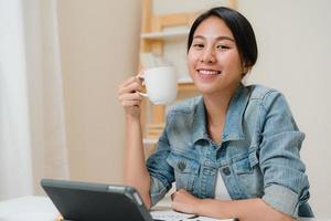 jonge aziatische vrouw die werkt met behulp van tablet die sociale media controleert en koffie drinkt terwijl ze ontspant op het bureau in de woonkamer thuis. genieten van tijd thuis concept. foto