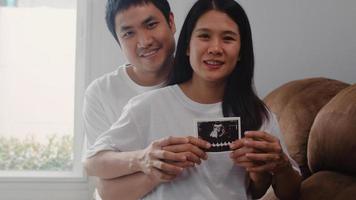 jonge Aziatische zwangere paar show en op zoek naar echografie foto baby in buik. mama en papa voelen zich gelukkig glimlachend vredig terwijl ze zorgen voor een kind dat op de bank ligt in de woonkamer thuis concept.