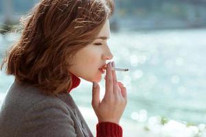 mooie stijlvolle trieste stressvolle jonge vrouw in trui en jas rokende sigaret in de buurt van de rivier foto