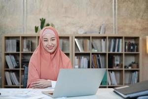 portret van de mooie oprichter van het startende bedrijf die een islamitische vrouwelijke persoon met hijab is, glimlacht en kijkt naar de camera in een kleine kantoorwerkplek, werkend met een laptop op een wit bureau, een boekenplank achter. foto