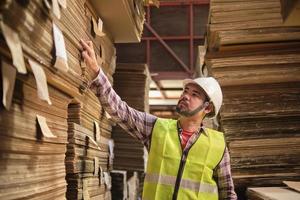 Aziatische mannelijke werknemer in veiligheidsuniform en helm, supervisor inspecteert verpakkingsvoorraadorder voor logistiek in fabrieksmagazijn, stapels stapelpapierfabricage, recyclingproductie-industrie. foto