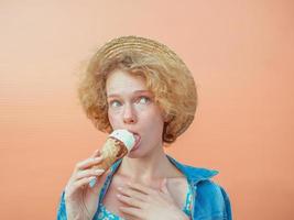jonge krullende roodharige vrouw in stro hoed, blauwe zomerjurk en jeans jasje eten van ijs op beige achtergrond. plezier, zomer, mode, jeugdconcept foto